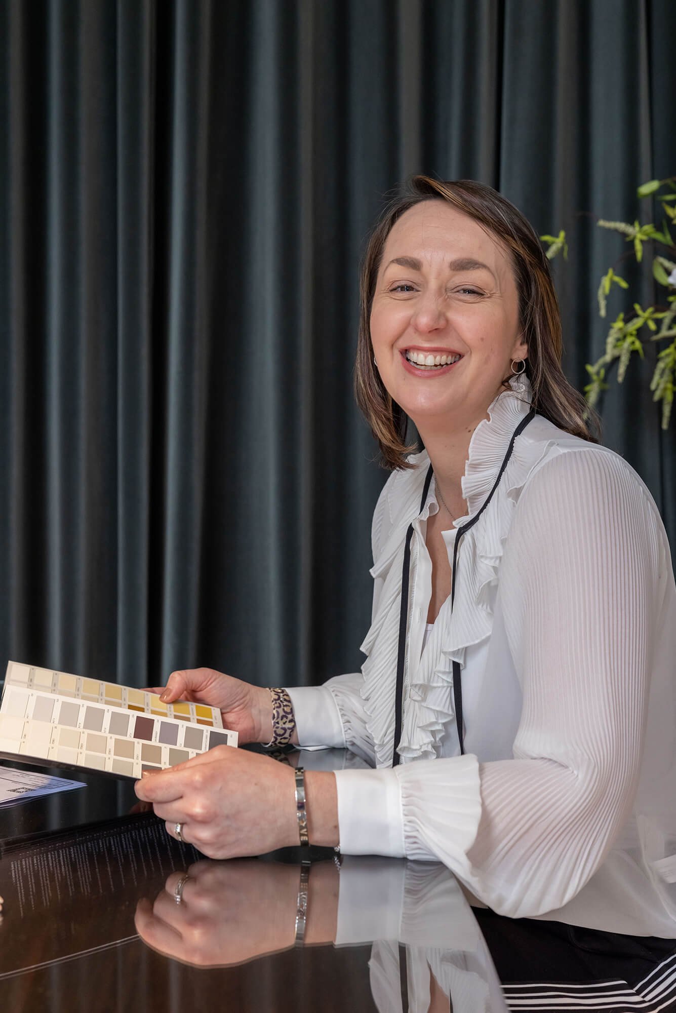 Professional interior designer smiles while holding color swatches, seated at a reflective table, ready for design.