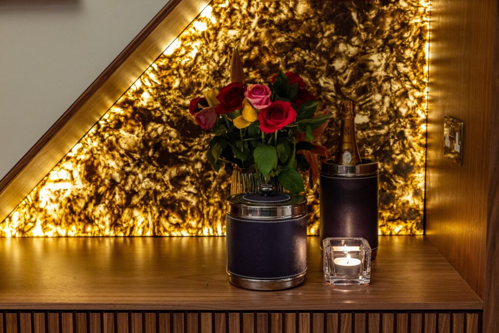 Stylish interior with a dramatic backlit amber onyx wall, a wood shelf displaying a vase of roses, champagne, and a candle.