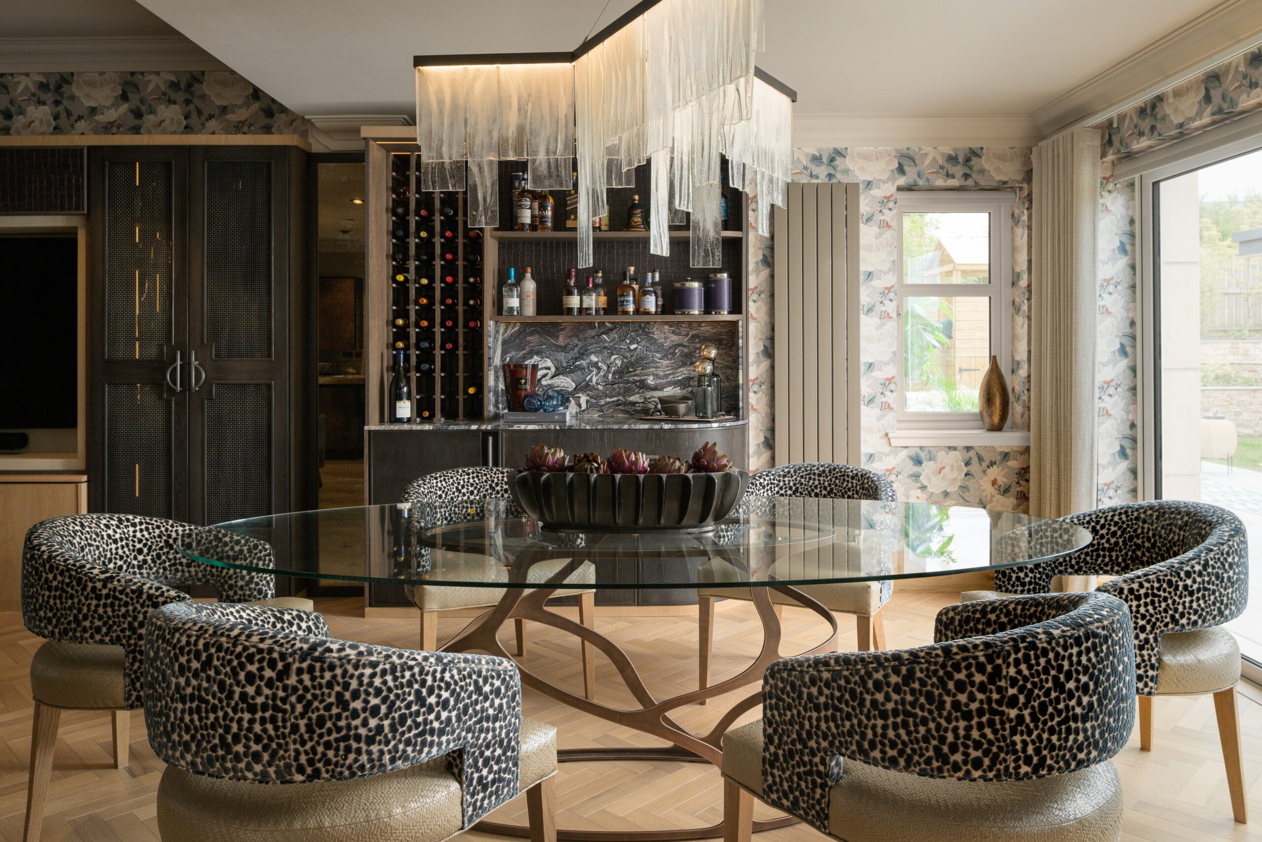 Elegant dining room: oval glass table, 6 leopard print chairs, modern chandelier, built-in bar, floral wallpaper, wood floor.