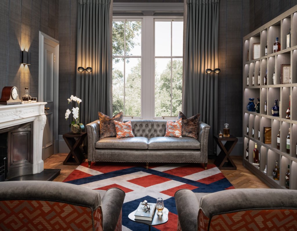 Sophisticated study with grey tufted sofa, colorful geometric rug, ornate fireplace, illuminated liquor cabinet, and armchairs.