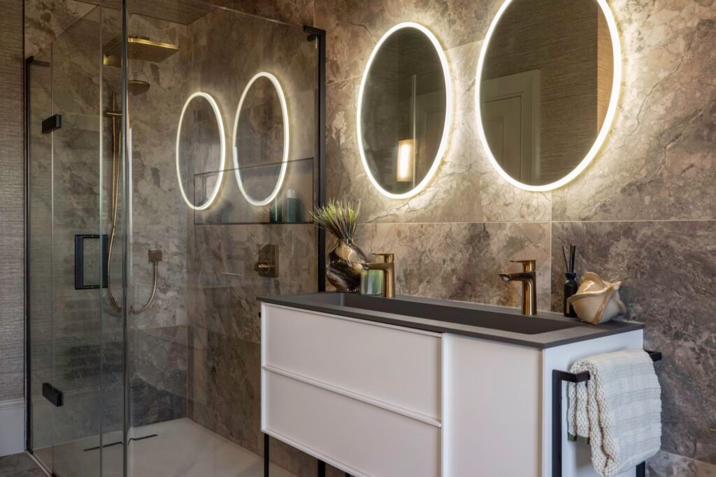 Modern luxury bathroom: double vanity, gold fixtures, backlit round mirrors, gray marble tiles, and glass shower.