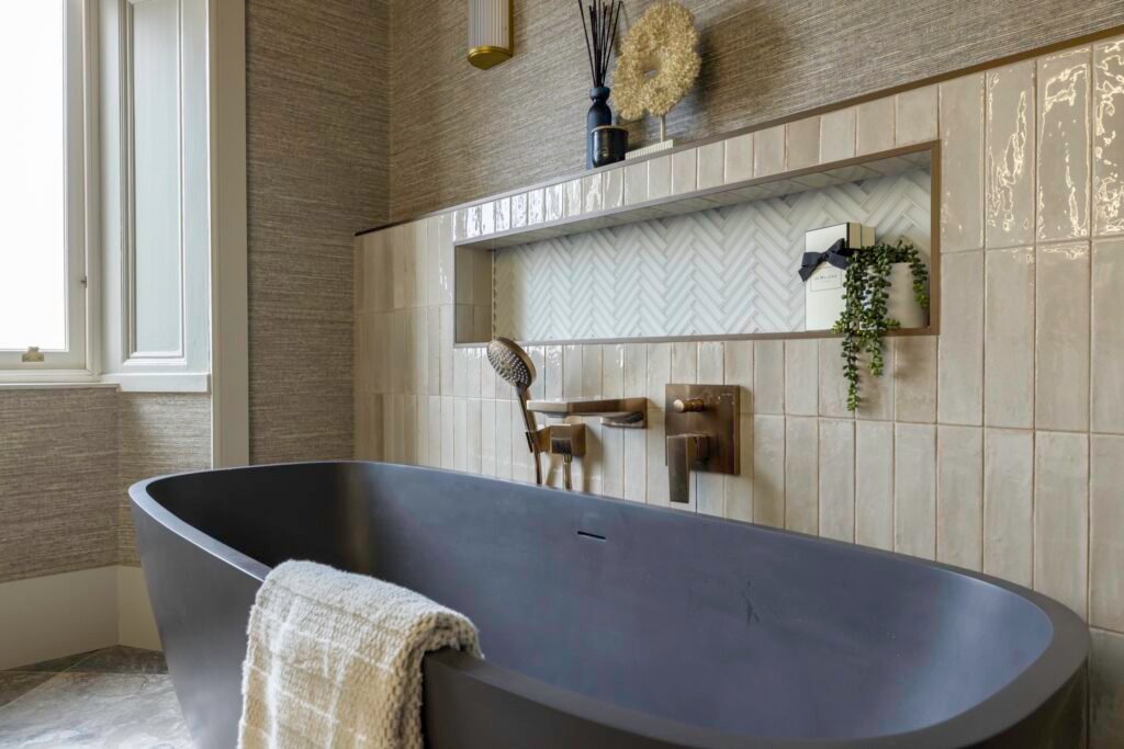 Modern bathroom with dark freestanding tub, cream textured tiles, bronze fixtures, woven wallpaper, and a herringbone niche.