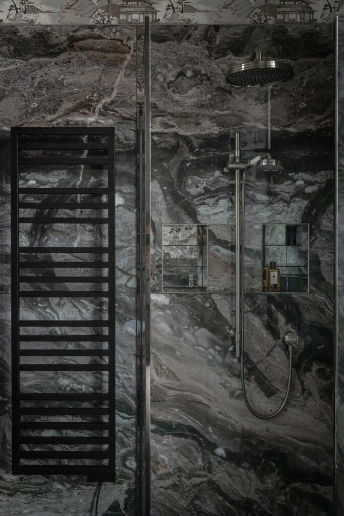 Luxurious shower with dark veined marble, rain shower head, handheld, black towel warmer, recessed shelves, and scenic wallpaper border.