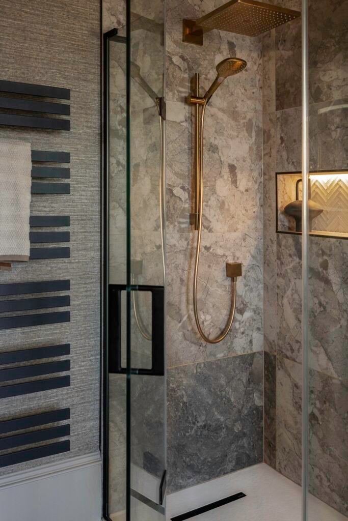 Modern bathroom shower with grey marble-effect walls, gold fixtures, illuminated niche, black towel warmer & textured wall.