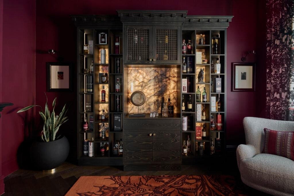 Dark wood built-in bar with backlit onyx, spirits collection, deep burgundy walls, textured armchair, and ornate rug.