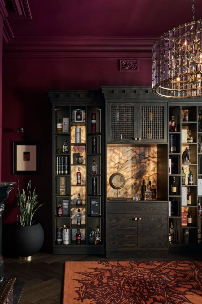 Luxurious home bar with deep plum walls, dark wood built-in shelving displaying liquor, backlit onyx, and a crystal chandelier.