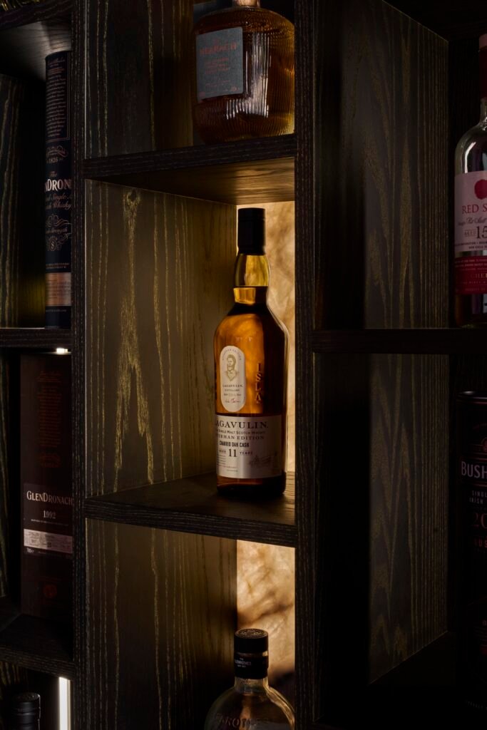 Elegant dark wood display with warm backlit shelves, filled with an assortment of premium whisky bottles.