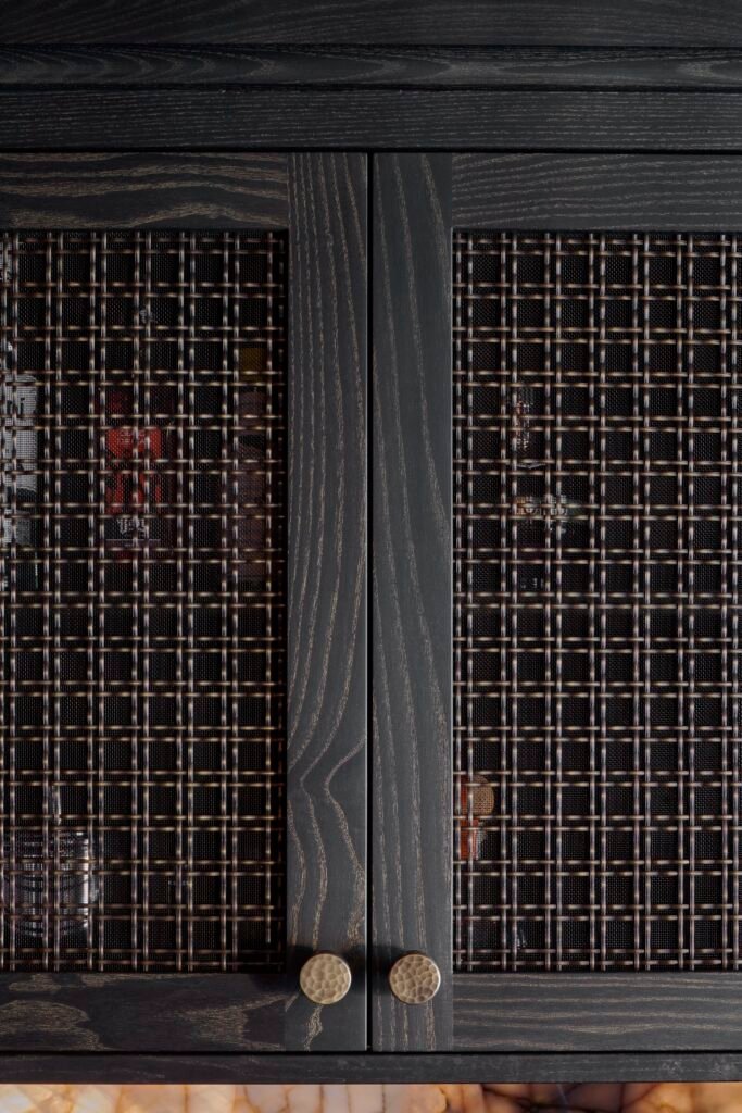 Dark wood cabinet doors with bronze woven metal mesh inserts and hammered brass knobs, above a backlit stone surface.