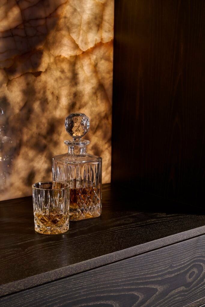 Sophisticated home bar featuring a crystal decanter and whiskey glass on dark wood, with a backlit onyx wall.