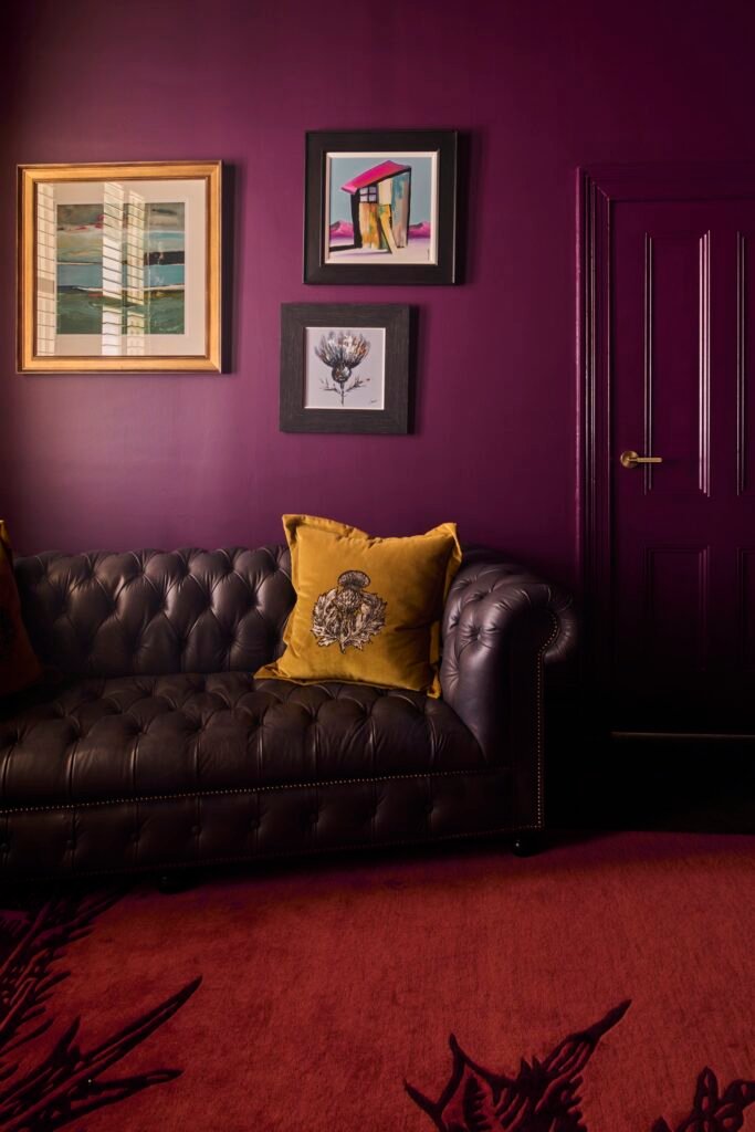Luxurious deep purple room with a tufted brown leather sofa, mustard yellow pillows, eclectic framed art, and a patterned red rug.