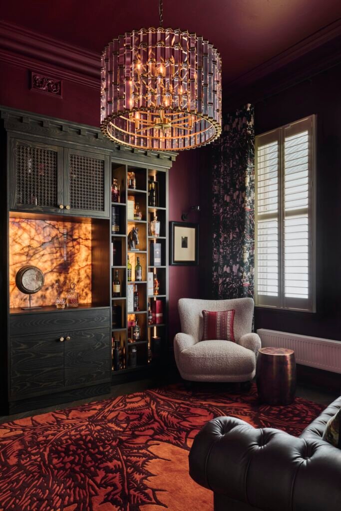 Opulent burgundy lounge with crystal chandelier, illuminated bar cabinet, patterned red rug, cream armchair, and leather sofa.