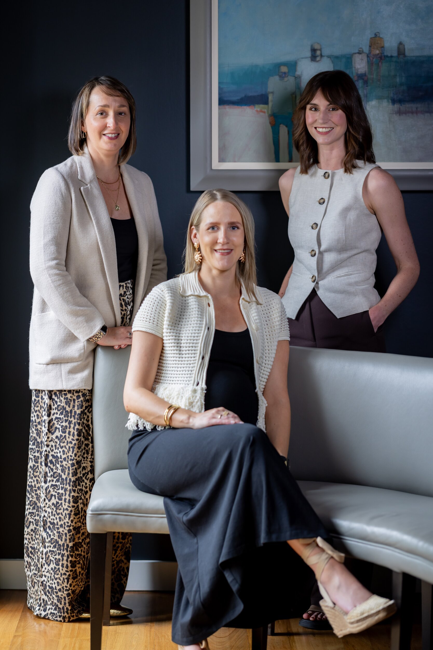 Three stylish women posing in a modern interior with dark walls, abstract art, and a grey upholstered bench.