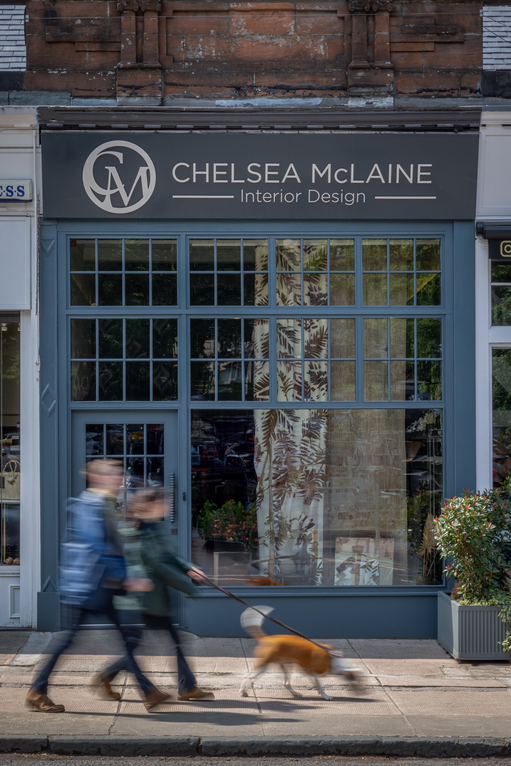 Chelsea McLaine Interior Design storefront with blue windows displaying textiles, people and dog blurred on the sunny sidewalk.