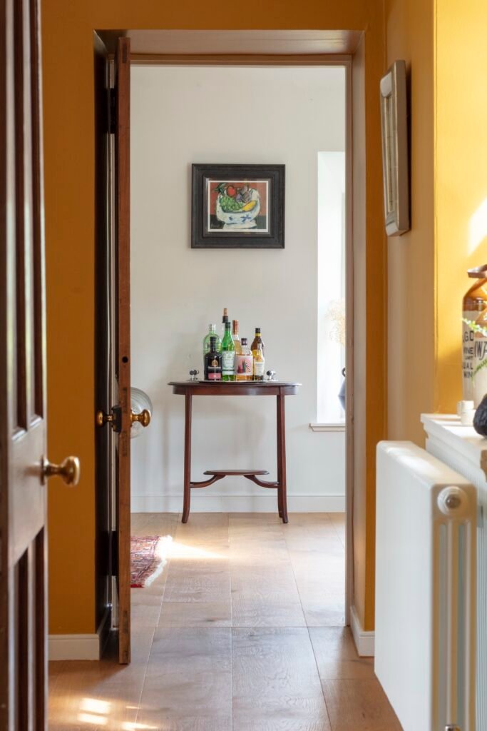View through an open doorway from a warm mustard hall into a white room with a console table of bottles and a fruit painting.
