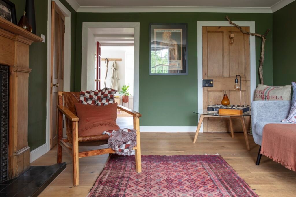 Cozy, eclectic living room with dark green walls, wood floors, a leather armchair, grey sofa, and a vibrant red patterned rug.