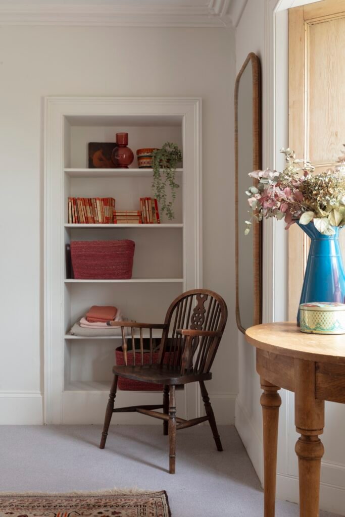 Charming interior with built-in white shelves, a vintage wooden chair, and a round side table with dried flowers and a mirror.
