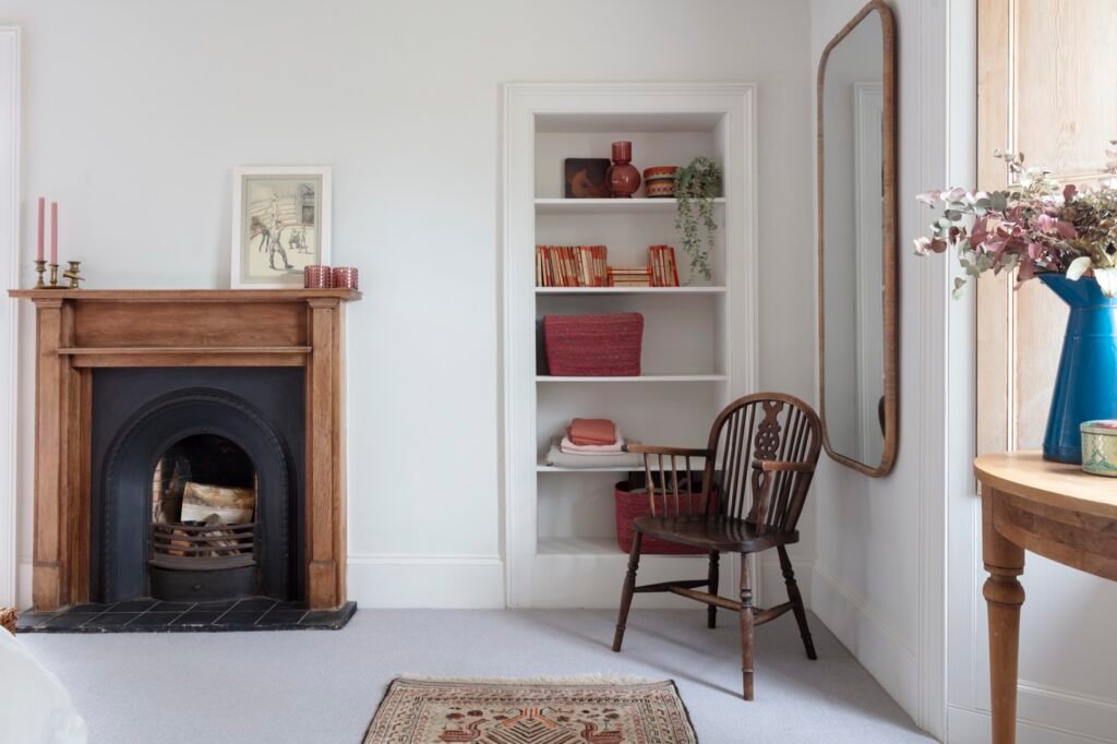 Bright room with wooden fireplace, white built-in shelves, dark wood Windsor chair, rattan mirror, and colorful accents.