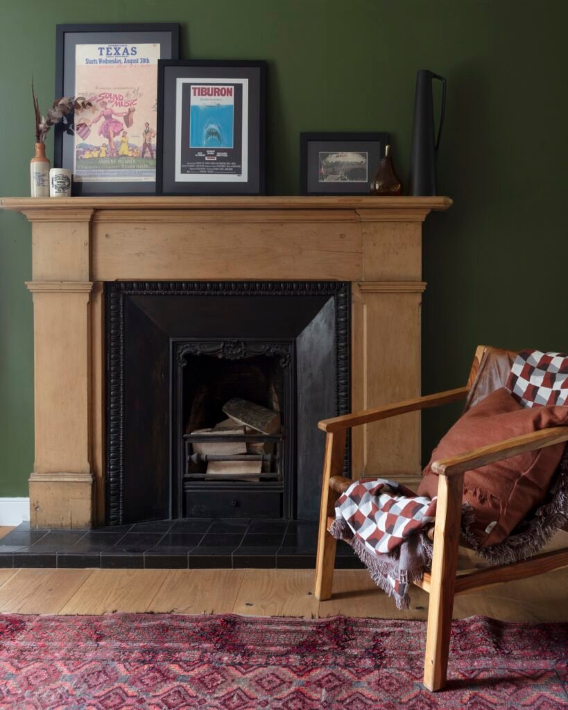 Warm living room with dark green walls, a wood fireplace, framed art, a leather armchair with throws, and a vibrant patterned rug.