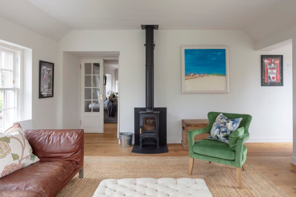 Bright, cozy living room with a wood-burning stove, brown leather sofa, green velvet armchair, white ottoman, and natural jute rug.
