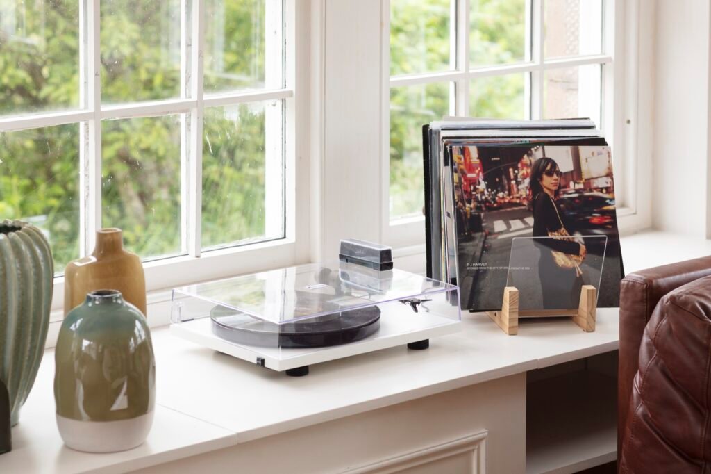 Cozy window nook with white turntable, stack of vinyl records, and displayed album cover. Vases and brown leather armchair.