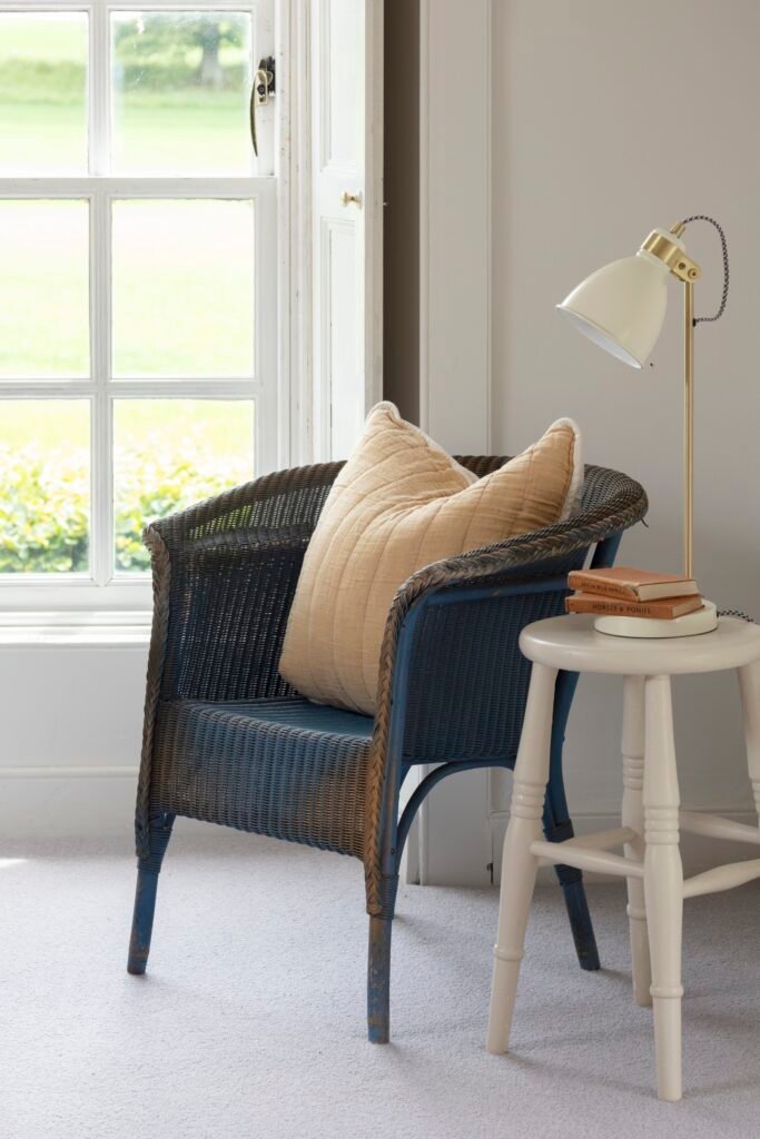 Cozy reading nook with distressed blue wicker armchair, tan pillow, cream lamp, books on a white stool, by a bright window.