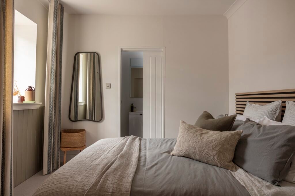 Serene neutral bedroom featuring a grey bed, slatted headboard, patterned curtains, rounded mirror, and bathroom access.