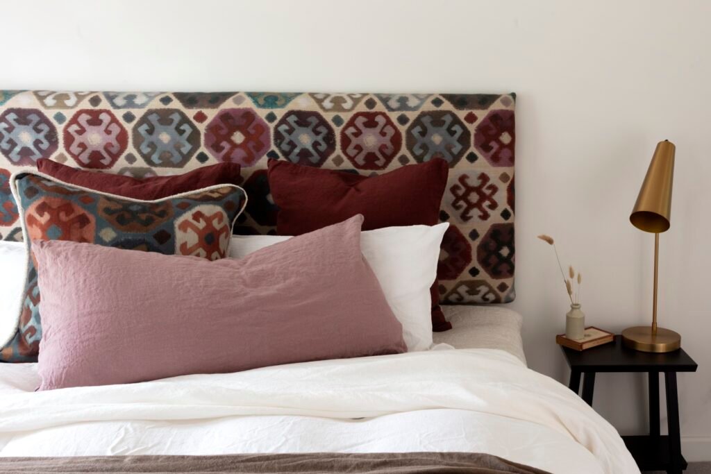 Eclectic bedroom with patterned ikat-style headboard, linen bedding, red & pink pillows, brass lamp on a black nightstand.