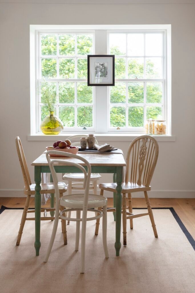 Farmhouse dining space with a distressed green table, varied wooden chairs, and bright window overlooking lush green trees.