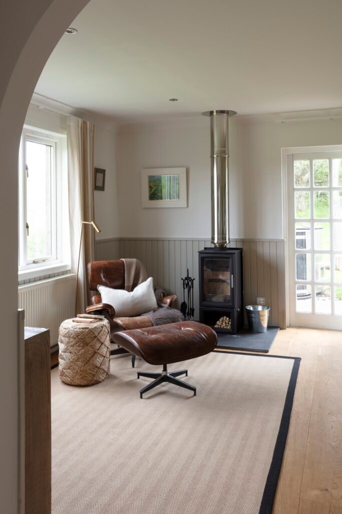 Cozy lounge area featuring a brown leather armchair, modern wood-burning stove, beige herringbone rug, and natural light.