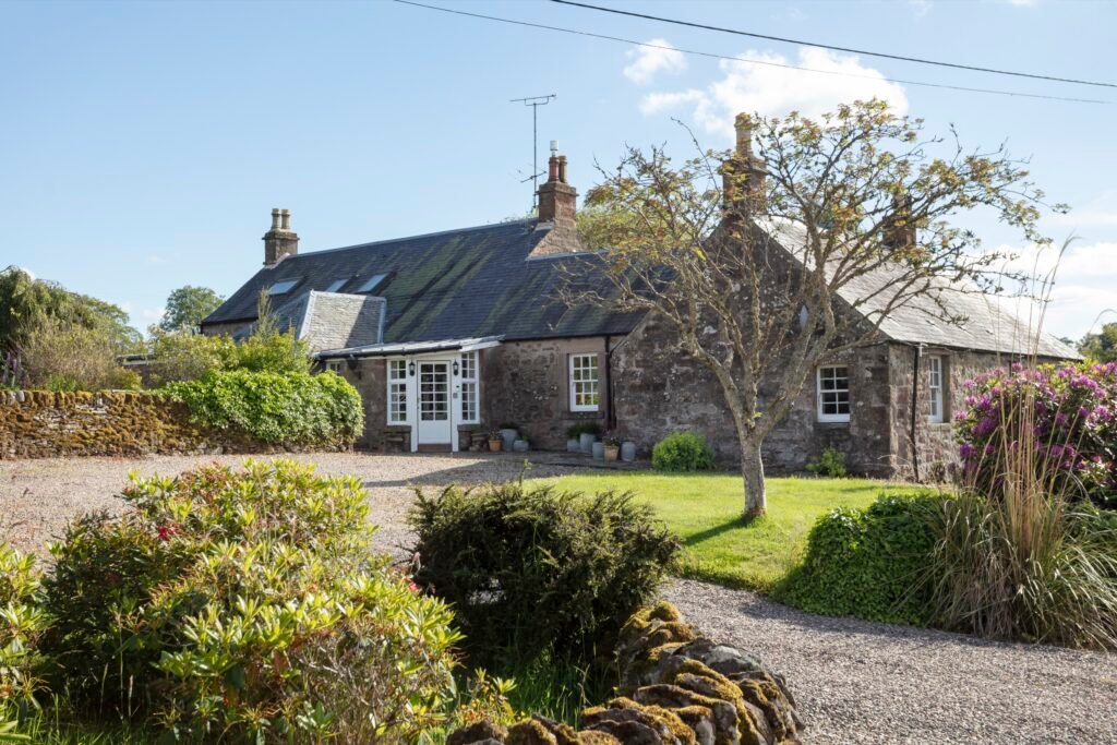 Traditional stone cottage with a slate roof, white windows and door, gravel drive, green lawn, and flourishing garden.