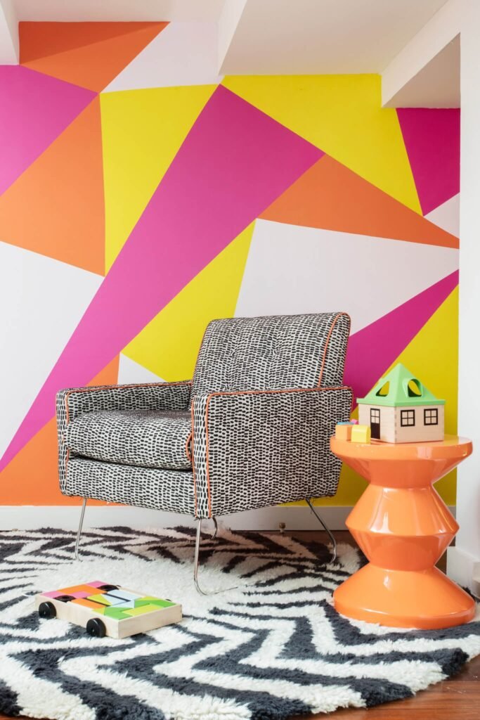 Playful room with bold pink, orange, yellow geometric wall, patterned armchair, orange side table, zebra rug, & wooden toys.