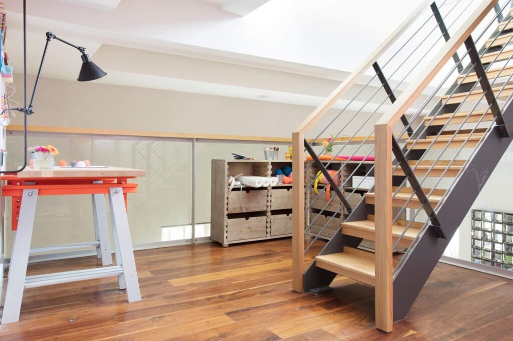 Modern creative workspace with an orange desk, craft storage, warm wood floors, and a sleek metal and wood staircase.