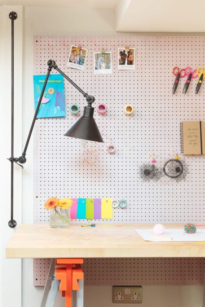 Vibrant craft workspace with wooden desk, pink pegboard wall holding art supplies, personal photos, and an industrial lamp.