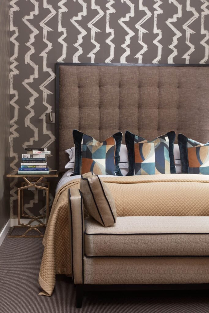 Modern bedroom with a brown tufted headboard, geometric blue-brown pillows, abstract zigzag wallpaper, and a patterned bench.