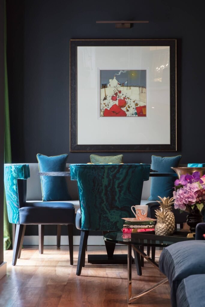 Dark blue room with a vibrant painting, patterned teal and blue chairs, and a coffee table with a golden pineapple and flowers.