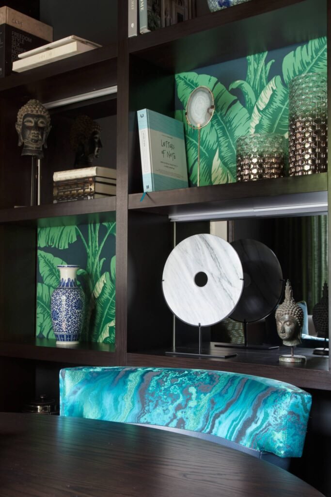 Dark wood shelves showcasing eclectic decor: tropical leaf wallpaper, Buddha heads, books, marble art, and a vibrant teal chair.