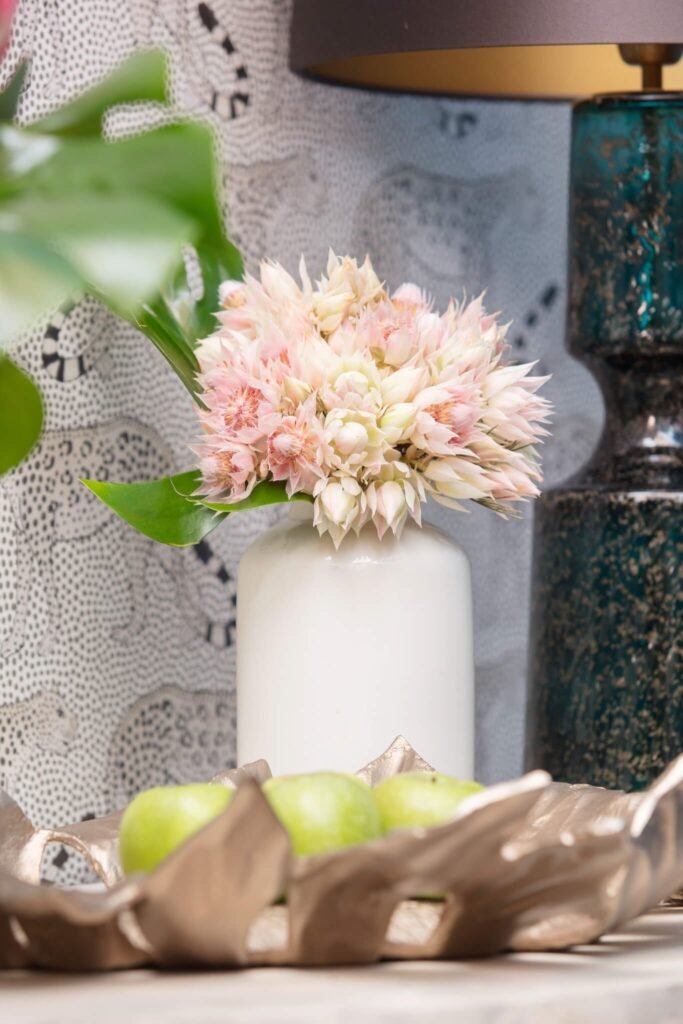 Interior design vignette with pink flowers in white vase, green apples in gold bowl, patterned wallpaper, and teal lamp.