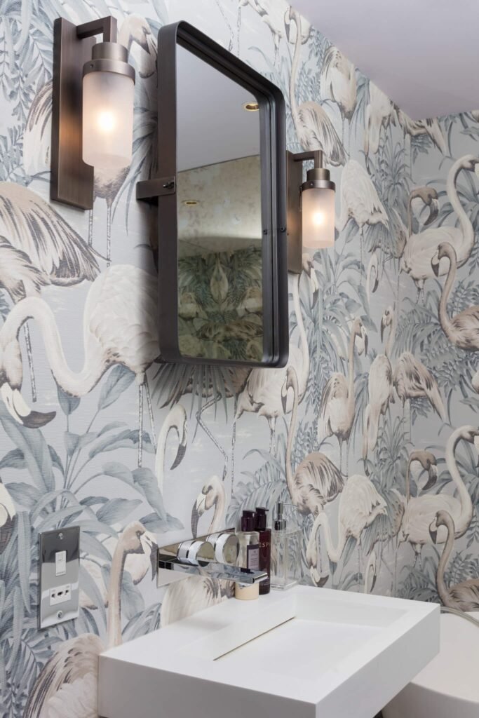 Stylish powder room with elegant tropical flamingo wallpaper, dark bronze framed mirror, matching sconces, and a white rectangular sink.