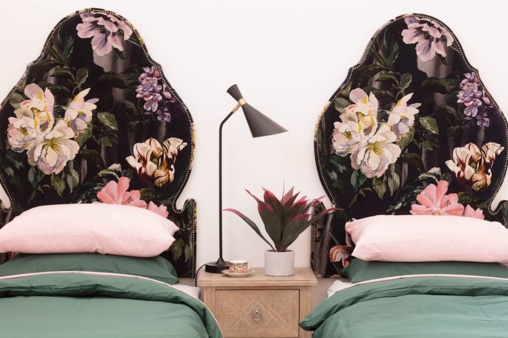 Twin beds with ornate dark floral upholstered headboards, pink and green bedding, a wooden nightstand, and a modern black lamp.