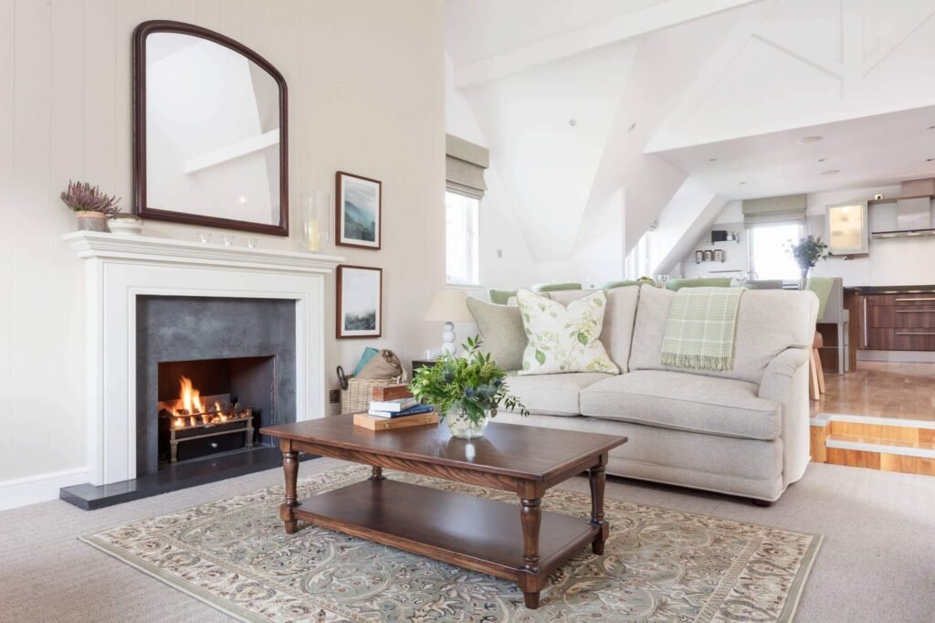 Cozy open-plan living room with a lit fireplace, arched mirror, beige sofa, wood coffee table, and connected modern kitchen.