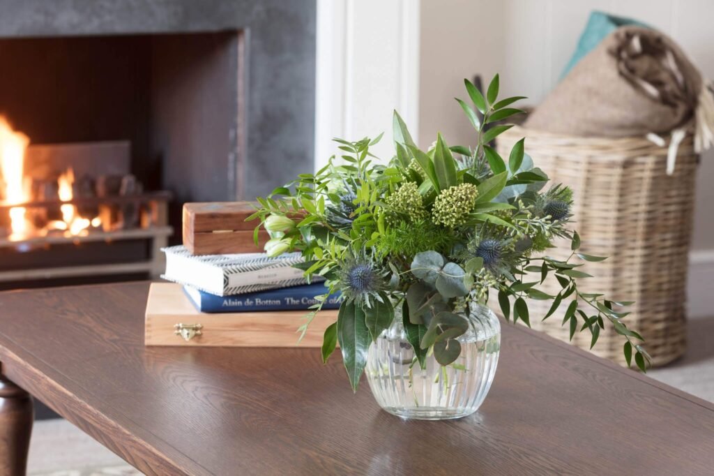 Vase with mixed greenery, blue thistles, and white buds on a dark wood coffee table by a lit fireplace in a cozy living room.
