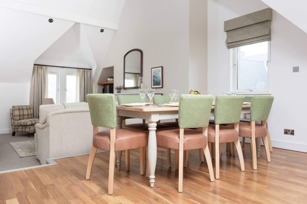 Modern open-plan living and dining room with sloped ceilings. Dining table set with green chairs, beige sofa, wood floor.