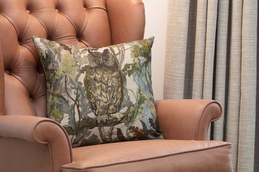 Tufted tan leather armchair featuring an owl & foliage patterned pillow, with neutral woven curtains in background.