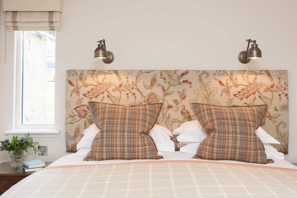 Cozy bedroom with a floral and bird upholstered headboard, plaid cushions, antique brass wall sconces, and checked bedding.