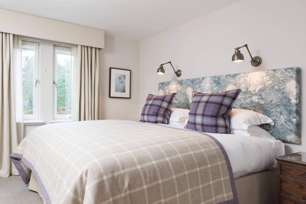 Bedroom with forest landscape headboard, purple plaid pillows, beige plaid throw, wall lights, and sunlit window.