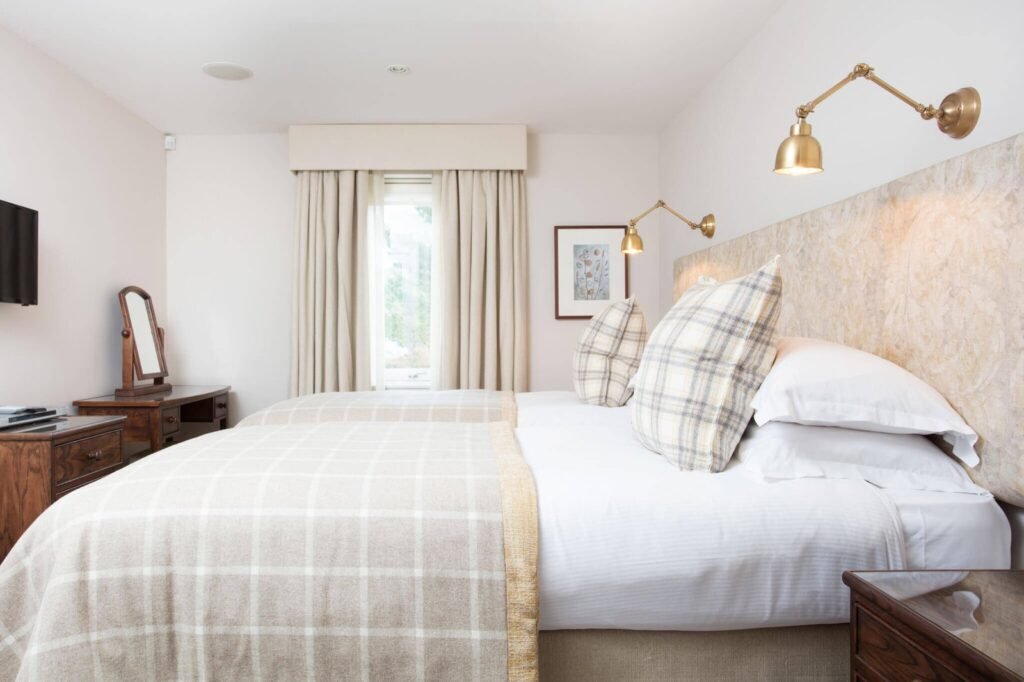 Elegant twin bedroom featuring neutral plaid throws, brass reading lamps, a wooden dresser, and cream curtains.