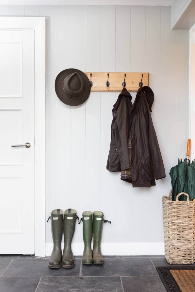 Mudroom with light grey wainscoting, wood coat rack with hat & jackets, green wellington boots, and wicker umbrella basket.