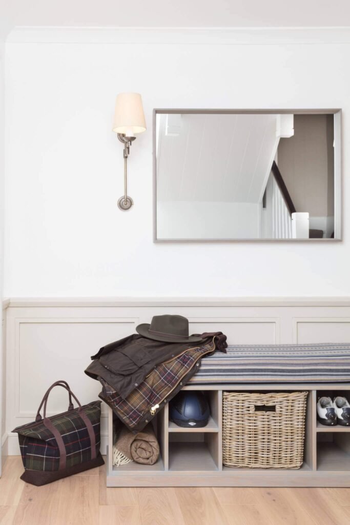 Tidy entryway with wooden storage bench, mirror, and wall light. Bench holds jacket, hat, helmet, shoes, basket. Plaid bag.