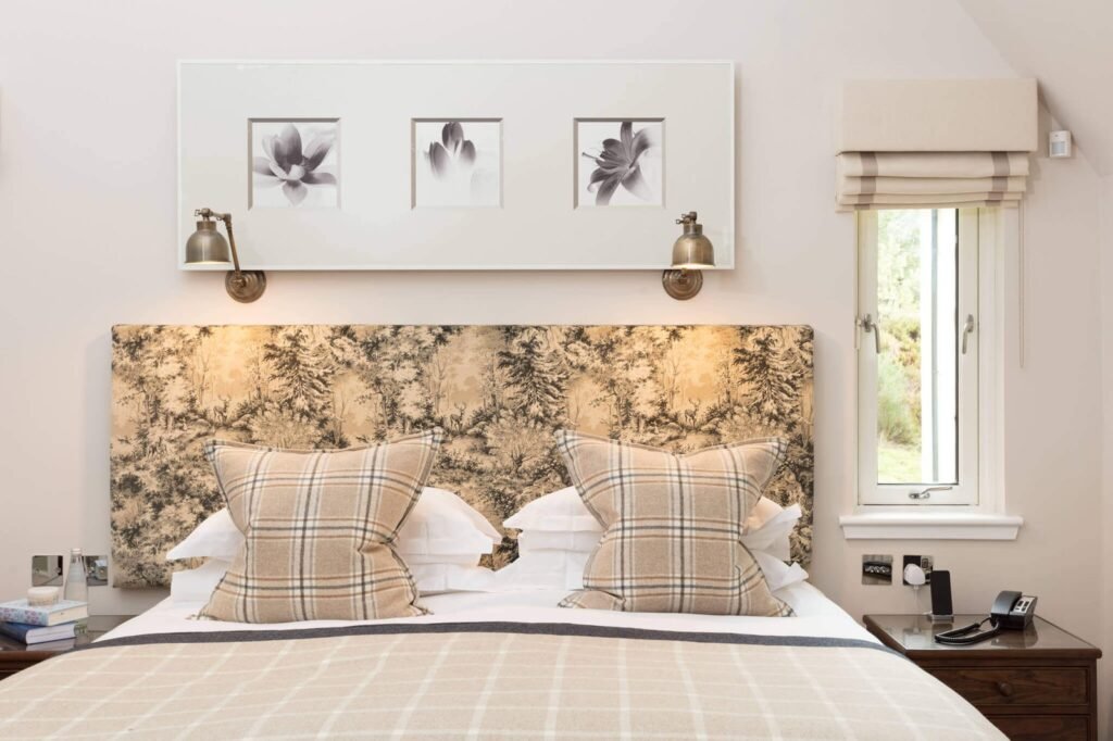 Cozy bedroom with a toile forest pattern headboard, plaid bedding, framed floral art, and antique brass reading lamps.
