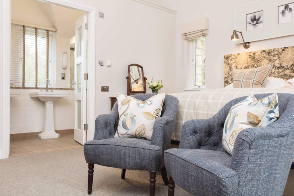 Elegant bedroom with comfy blue armchairs, floral cushions, a bed, and a bright ensuite bathroom with a pedestal sink.
